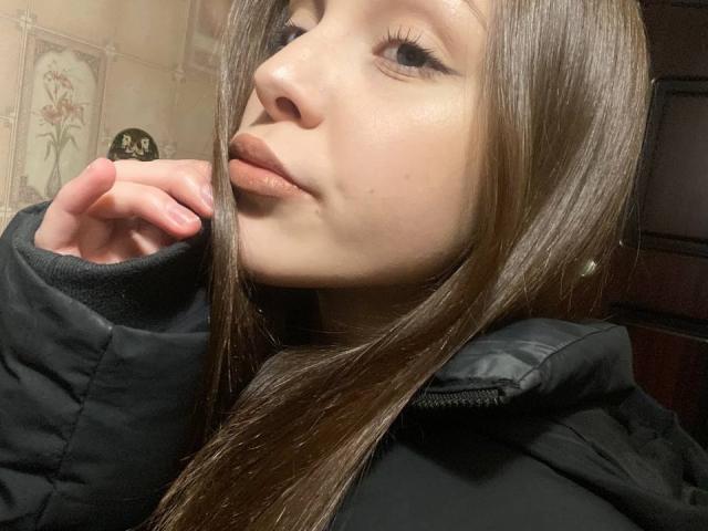 Danniette Snuggs met rechtbruin haar, licht open lippen, en een zwarte, opvallende jas, hand in de buurt van de mond, naar boven kijkend, bleke huid, subtiele make-up en lange wimpers..