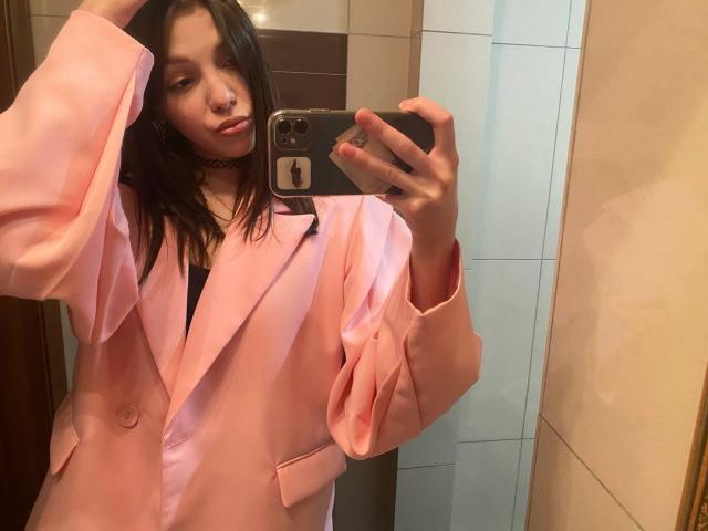 Danniette Snuggs, con cabello largo y negro, vestida con un blazer rosa sobre una blusa negra, tomando una selfie en un espejo, con un collar tipo "choker", sosteniendo un teléfono con una funda oscura..