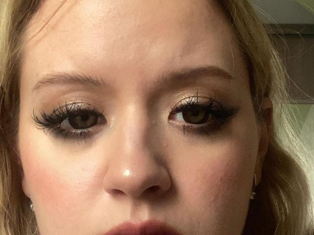 AureliaHavis's face, showing brown eyes with thick black eyelashes, light skin, and a slight sheen on her nose. Blonde hair frames her face. Her lips are closed.