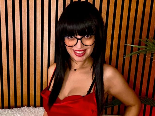 SofiaNoireX, com óculos pretos, blusa de cetim vermelha e alças de sutiã pretas, sorrindo com batom vermelho, cabelos longos pretos e um colar de coração, mostrando seus seios..