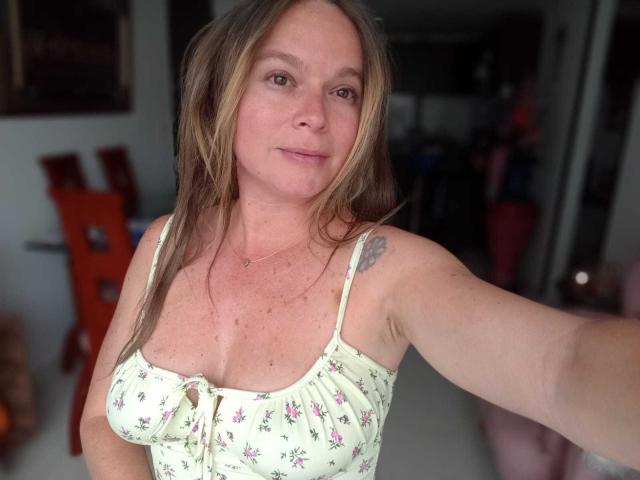 PamelaColin with light brown hair, small pink flower dress, visible tattoo on right shoulder, light skin, small boobs, slightly flushed, taking a selfie indoors.
