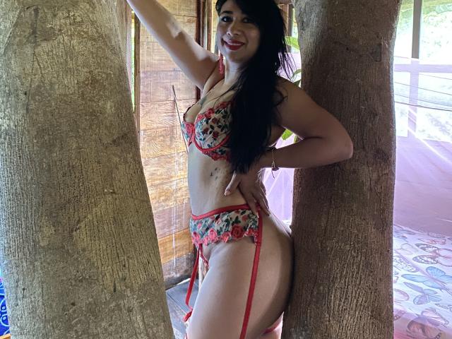 Karimelee stands between two trees, wearing red floral lingerie with garters, showing her curvy ass and small tits. She smiles, with long black hair, and red lipstick.