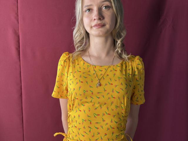 ChirquitaDagata con cabello rubio y ondulado, vestido amarillo con estampados rojos y verdes, mangas cortas, pequeños senos, collar, sonriendo, de pie frente a una tela roja..