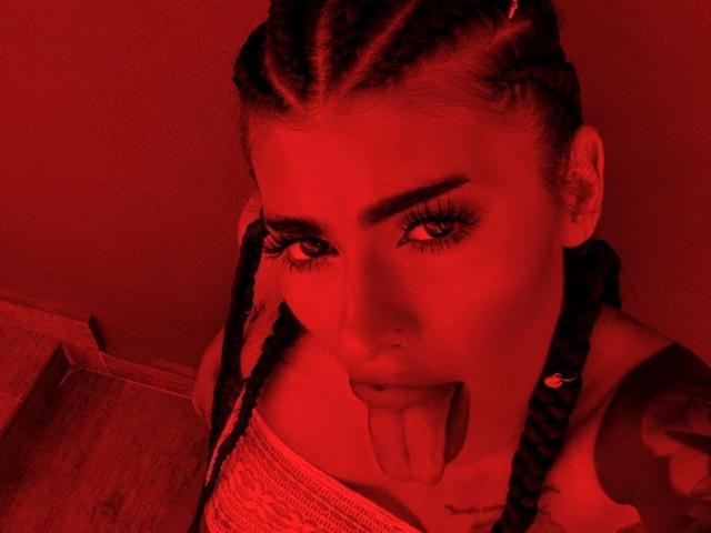 Red-lit selfie of DariaAmaris with braided hair, nose ring, and tongue out, showing off her tattoos and long eyelashes. Her tongue is sticking out playfully.