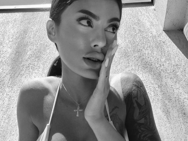 Black and white photo of DariaAmaris with dark hair in a ponytail, hand on cheek, cross necklace, tattoo on right arm, looking up, wearing a halter top, showing tits.