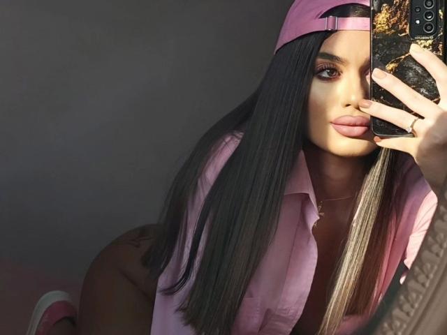DariaAmaris with long black hair, pink shirt, and pink cap, taking a mirror selfie, showing her ass and pink lips, holding a phone with a black case.