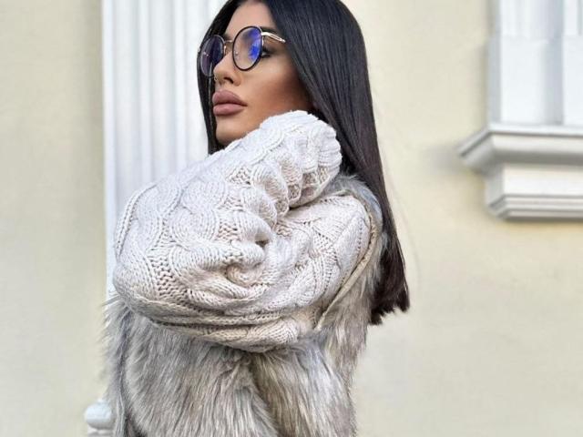 DariaAmaris with long black hair, wearing round glasses, a fluffy white knitted sweater, and a grey fur coat, looking over her shoulder.