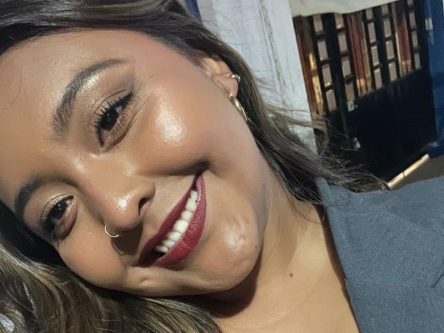 RushRosee's face, smiling with red lipstick, nose ring, and shiny skin, wearing a gray shirt, brown hair, and gold earrings. Boobs not visible.