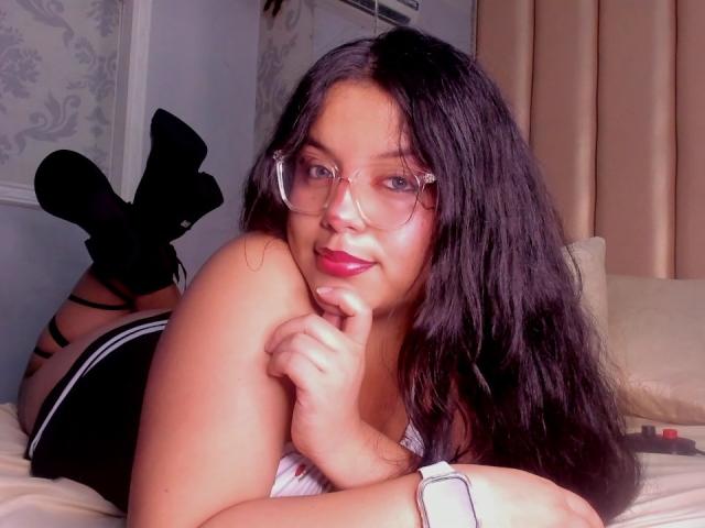 KonyBella with long black hair, glasses, pink lipstick, lying on bed, black socks, black-and-white top, smiling, hand on chin, legs up, white wristwatch, soft bedroom light.