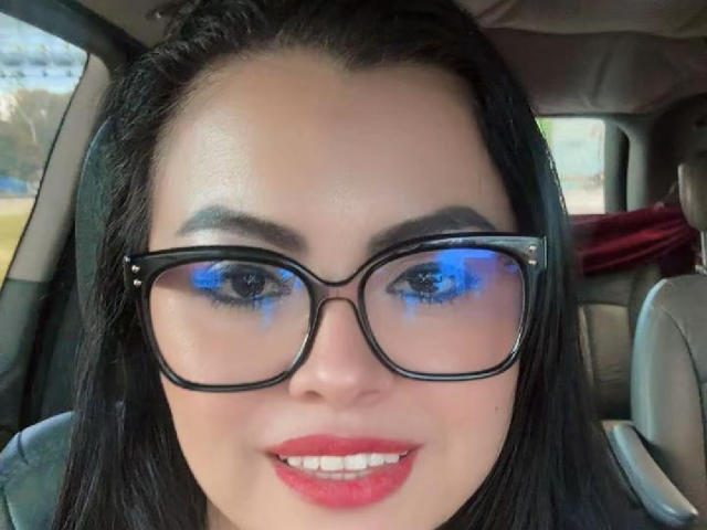 CharlotteDevereaux with black-framed glasses, blue reflections in glasses, red lipstick, and black hair, smiling in a car. Her tits are not visible.