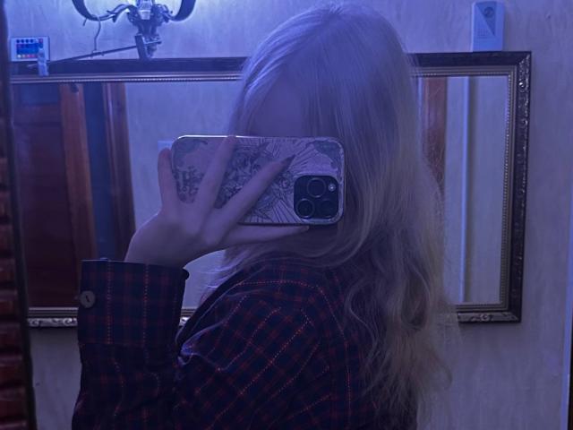 TiffaniMacandog with blonde hair, wearing a red plaid shirt, holding a phone with a white and black patterned case, taking a mirror selfie, covering her face.