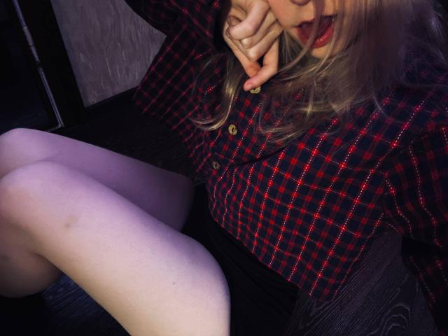 TiffaniMacandog in a red-black checkered shirt, biting her finger, with her legs up, showing pale skin and a small bruise on her thigh. Her red lipstick matches her shirt.