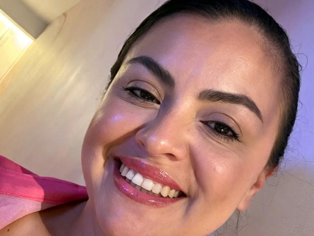 CharlotteDevereaux's face, smiling with glossy pink lips, white teeth, and brown eyes. She has dark eyebrows, shiny skin, and a pink shirt partially visible on her right.