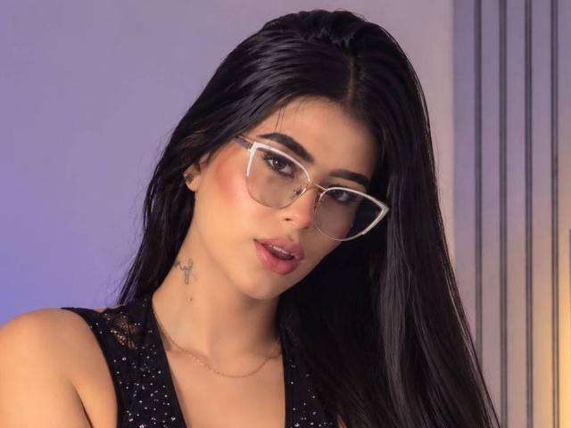 AmilyBella with long black hair, wearing clear glasses, black sparkly top, and a small cross tattoo on her neck, looking at the camera with slightly parted pink lips.