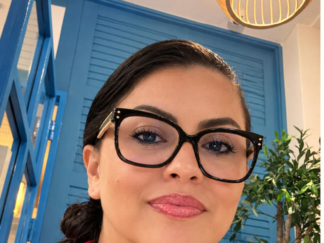 CharlotteDevereaux with shiny pink lipstick, black glasses, and dark brown hair in a bun, smiling at the camera. Blue shutters in the background. Her ass is not visible.