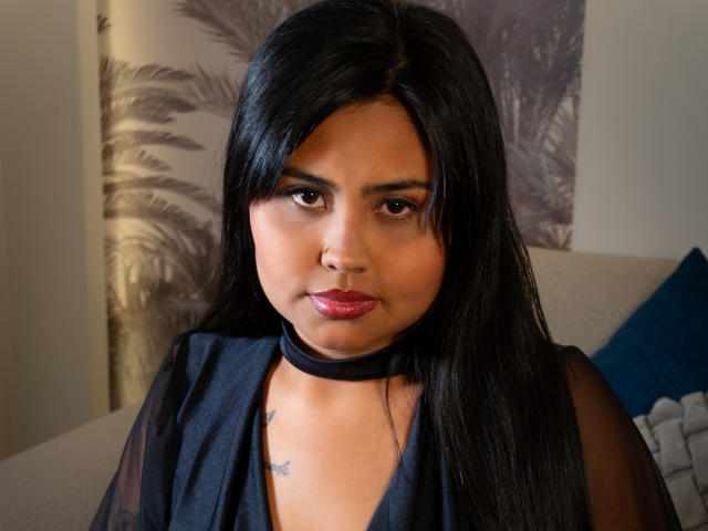 LuceroBe has dark, straight hair, brown eyes, and red lipstick. She's wearing a black choker and sheer top, with small tattoos on her chest. Her skin is tan.