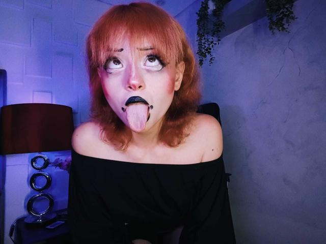 ViolettClay with red hair, black off-shoulder top, and black lipstick, sticking out her tongue with eyes wide, looking up. Her tongue is pink, and her eyes are exaggerated.