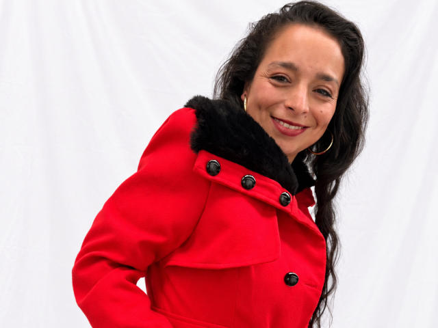 StangelarRivera, con cabello largo y negro, sonriendo, vestida con un abrigo rojo con cuello y botones de piel negra, fondo blanco, con aros grandes, mirando ligeramente hacia abajo, sonriendo..