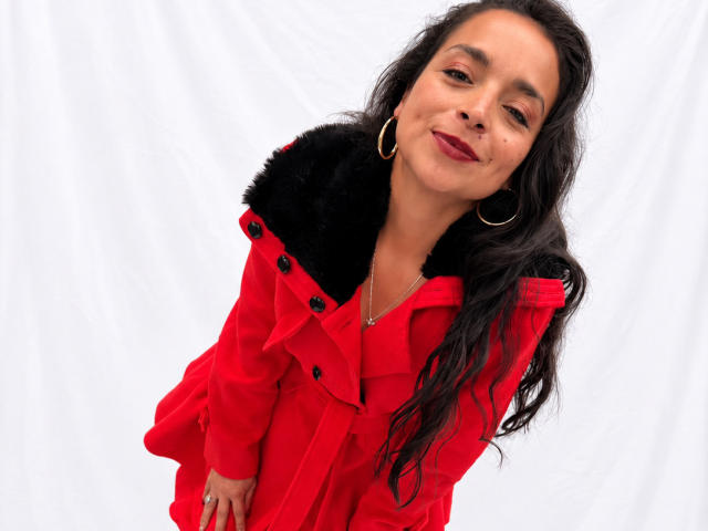 StangelarRivera with long black hair, red coat with black fur collar, gold hoop earrings, and red lipstick, smiling slightly, showing small tits, against a white background.