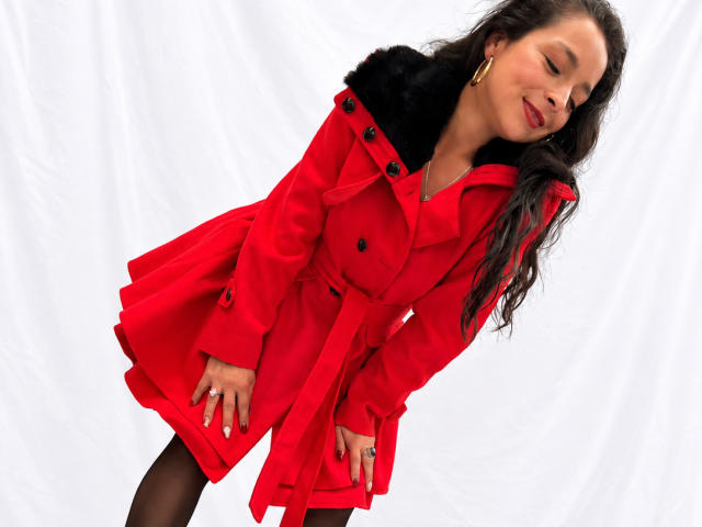 StangelarRivera with long black hair, red coat, black fur collar, black stockings, gold earrings, leaning forward, smiling, hands on coat, white background, red nails, looking cute.