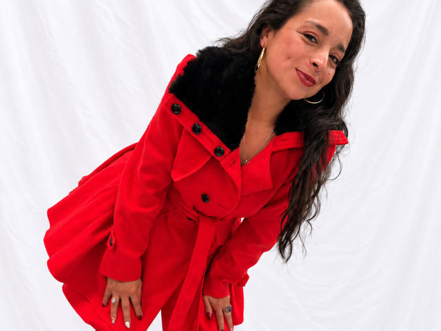 StangelarRivera, con cabello largo y oscuro, abrigo rojo, cuello de piel negra, aretes de aro dorados, inclinada hacia adelante, sonriendo, lápiz labial rojo, fondo blanco, manos sobre el abrigo, anillo en la mano derecha..