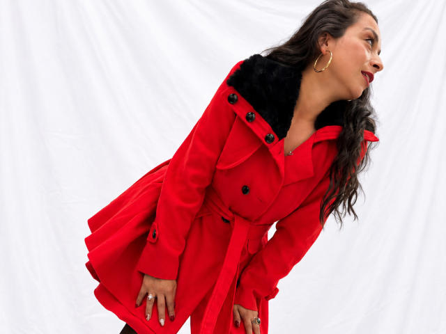 StangelarRivera indossa un cappotto rosso con un colletto di pelliccia nera, è china in avanti, porta orecchini a cerchio dorati, rossetto rosso e anelli, ha lunghi capelli castani ondulati e guarda di lato..