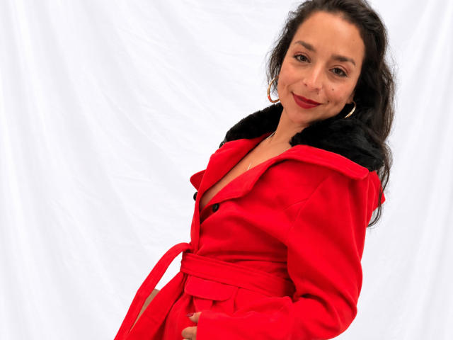 StangelarRivera with red coat, black fur collar, and red belt, smiling, red lipstick, dark wavy hair, white background, wearing hoop earrings, slight tilt, confident pose.