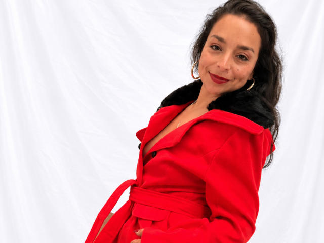 StangelarRivera con cappotto rosso, colletto di pelliccia nera e cintura rossa, sorridente, capelli scuri ondulati, rossetto rosso, orecchini a cerchio dorati, sfondo bianco, in piedi, rivolta verso la fotocamera..