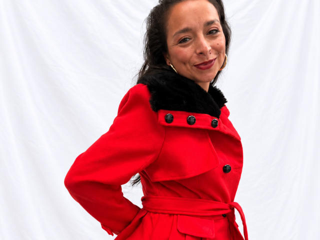 StangelarRivera, con seni di media grandezza, indossa un cappotto rosso con colletto in pelliccia nera, bottoni neri, e ha le mani sui fianchi, sorridendo. Ha capelli scuri, pelle chiara, orecchini a cerchio e uno sfondo bianco..