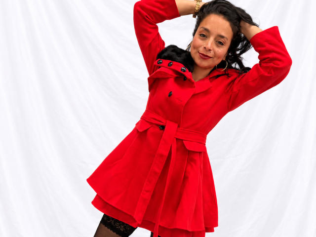 StangelarRivera with arms raised, wearing a red dress, black lace stockings, and hoop earrings, smiling at the camera. She has medium tits and dark wavy hair.