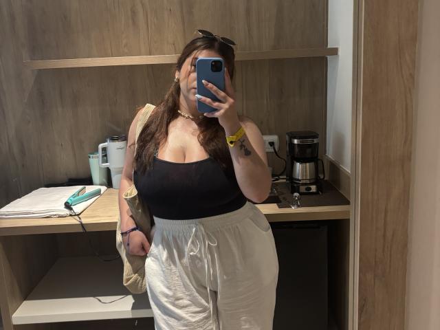VenusxMusa with large boobs, black strapless top, white loose pants, brown hair, blue phone, gold necklace, tattoo on left forearm, mirror selfie in a wooden room.