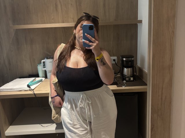 VenusxMusa with large tits, wearing a black strapless top, white loose pants, holding a phone in front of her face, in a wood-paneled room with a coffee maker.