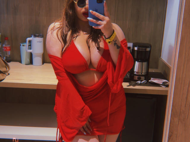 VenusxMusa with huge tits in a red, cut-out dress, off-shoulder sleeves, holding a phone in front of her face, standing in a room, wearing sunglasses.