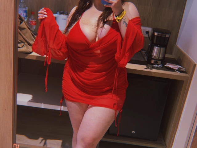 VenusxMusa in a red dress with deep cleavage, showing her tits, holding a red ball, mirror selfie, coffee maker in background, wooden shelf, yellow bracelet on wrist.