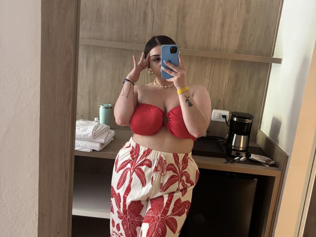 VenusxMusa with large boobs, red strapless top, and white floral pants, taking a mirror selfie, holding a blue phone, in a room with a coffee maker and towels.