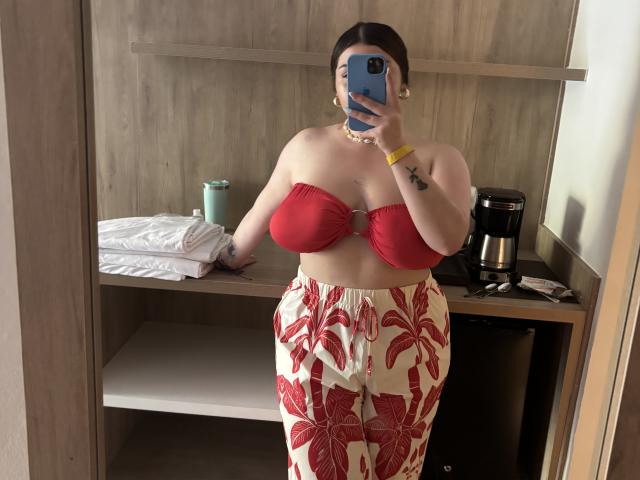 VenusxMusa with red strapless top, showing big boobs, and red floral pants, holding a phone in front of her face, standing in a room with wooden shelves.