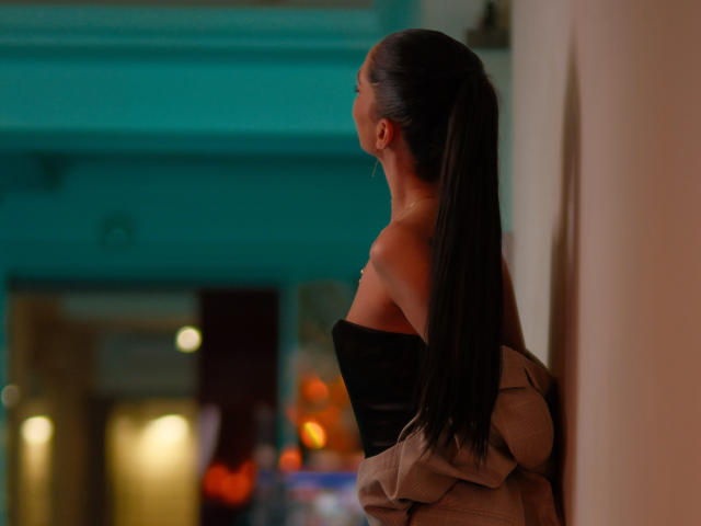 EllyLynne with long black ponytail, off-shoulder black top, and beige jacket, standing against a wall, looking to the side, in dim indoor lighting.