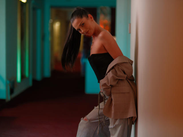 EllyLynne with dark ponytail, black strapless top, beige jacket off-shoulder, leaning against wall, looking down, red carpet, blue-green walls, her tits slightly visible, slim, sexy.