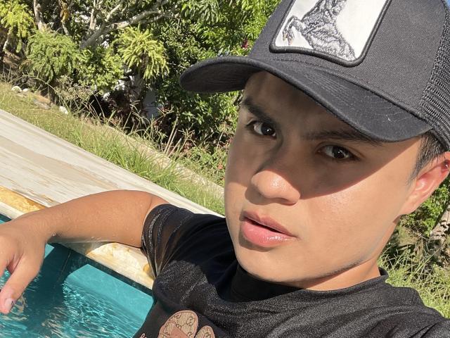 ThiagoAriass, wearing a black cap with a graphic, leans on a pool's edge, brown eyes looking at the camera, black shirt, short brown hair, slight tan, sunlit.