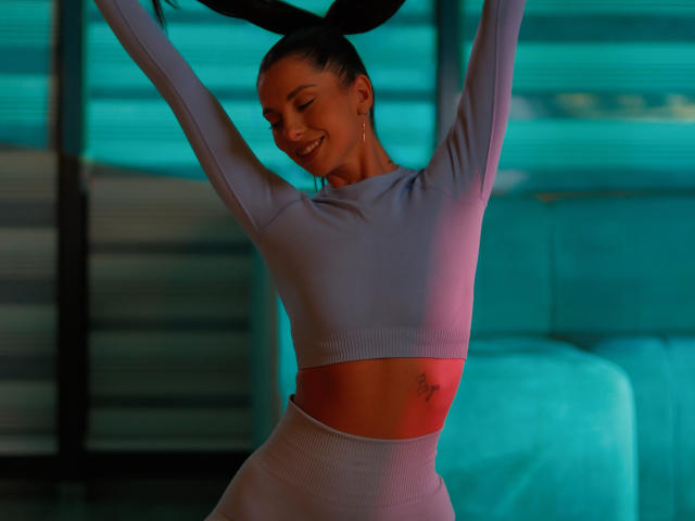 EllyLynne with dark hair in bunny ears, smiling, arms raised, wearing a light purple crop top and high-waisted pants, showing a small tattoo under her right breast.