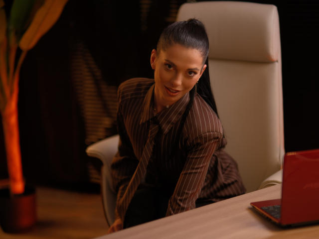 EllyLynne with ponytail, brown pinstripe suit, leaning on white office chair, smiling, laptop on desk, red cover, wooden floor, orange plant on left, dark background.
