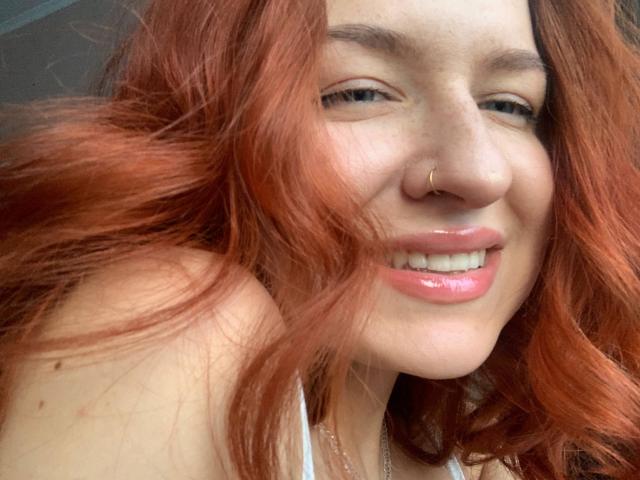 Ernestina Mcintrye tiene el pelo rojo y ondulado, un piercing en la nariz y labios rosados y brillantes. Sonríe a la cámara, y se pueden ver ligeramente sus pechos a través de una blusa blanca..