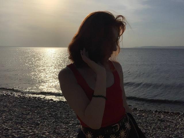 ErnestinaMcintrye with red hair, wearing a red tank top and black skirt with sun patterns, holding phone to ear, standing on pebbled beach, sunlight reflecting on water.