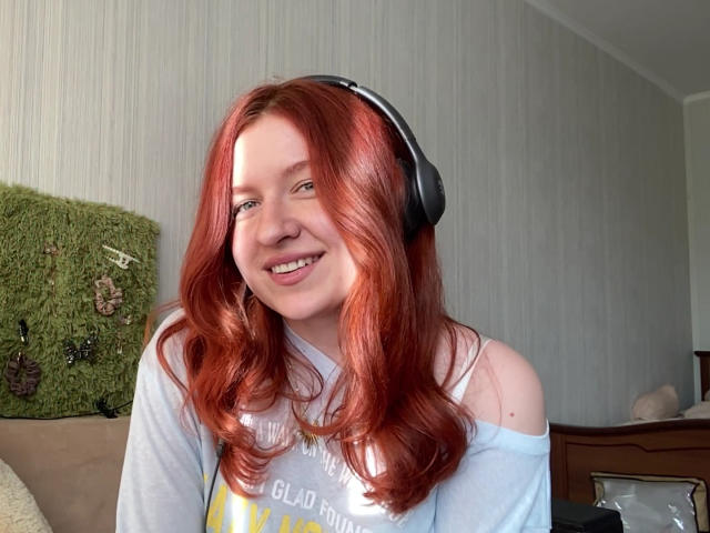 ErnestinaMcintrye with red hair, smiling, wearing headphones, light blue off-shoulder shirt, and a white top, showing her tits slightly. She has a cute face and looks happy.