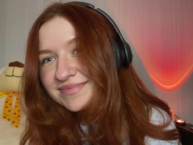 Ernestina Mcintrye, com cabelo ruivo, usando fones de ouvido, sorrindo, tem pele clara e lábios rosados, em um selfie, com um brinquedo de pelúcia e uma luz vermelha ao fundo..