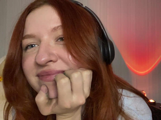 Ernestina Mcintrye hat rotes Haar, blaue Augen und lächelt, wobei sie ihre Hand unter das Kinn legt. Sie trägt schwarze Kopfhörer, sieht süß und entspannt aus..