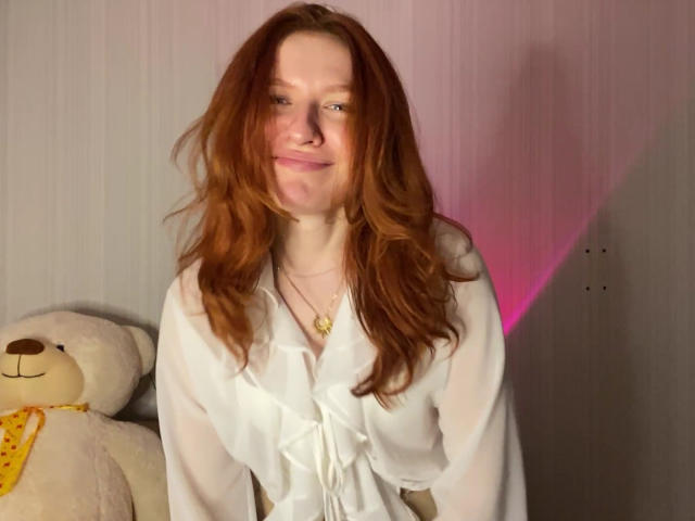 Ernestina Mcintrye, met rood haar, een witte blouse en een gouden ketting, glimlacht lichtjes en staat naast een witte teddybeer met een gele sjaal. Haar borsten zijn niet zichtbaar..