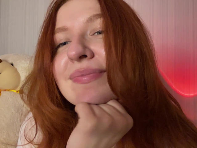 ErnestinaMcintrye met rood haar, volle, roze lippen, hand onder haar kin, die zachtjes glimlacht, lichte huid, blauwe ogen, en een schattige knuffelbeer aan de linkerkant..