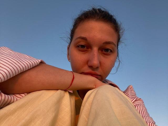 Sanella with her head resting on her knees, wearing a red-striped shirt and white pants, has a red bracelet on her wrist, under a clear blue sky.