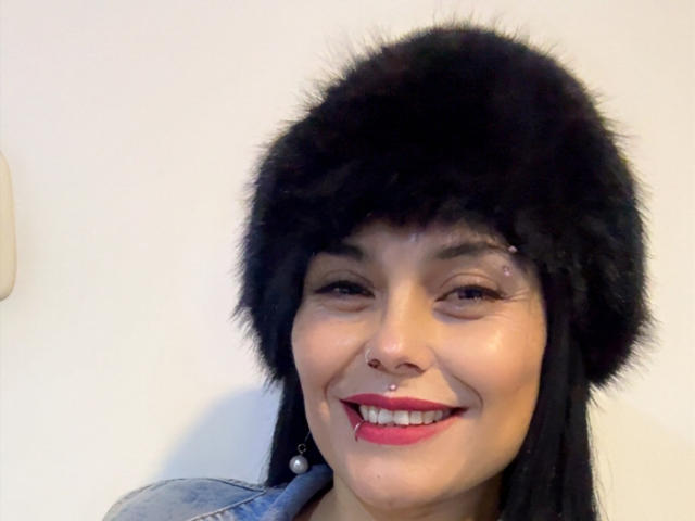 CharlotteCrofort with black fur hat, black hair, red lipstick, nose piercing, smiling, wearing denim jacket, white background, visible tear on right cheek, looking at camera.